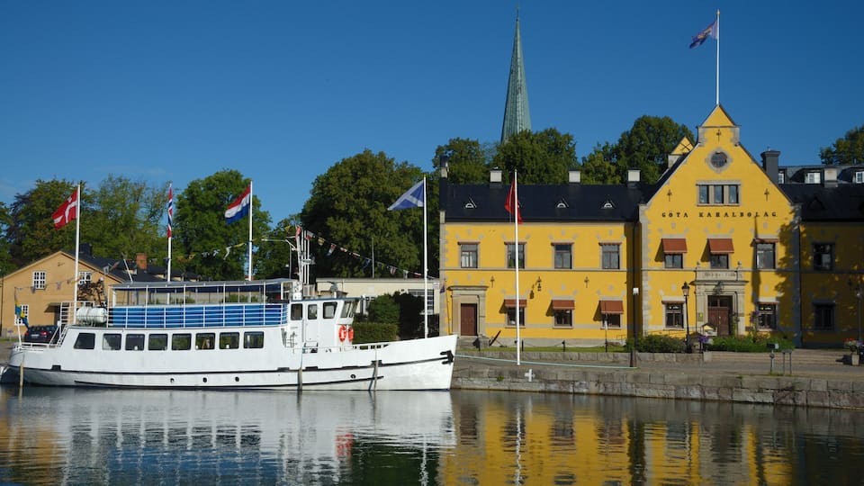 A white boat docked near a yellow building with flags, a church spire, and a calm water body reflecting the scene.