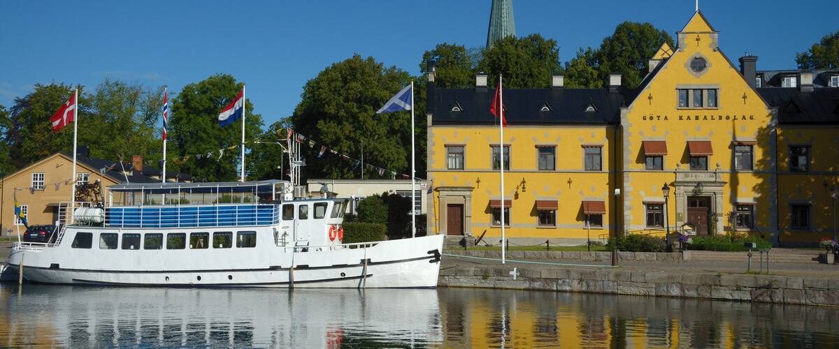 A white boat docked near a yellow building with flags, a church spire, and a calm water body reflecting the scene.