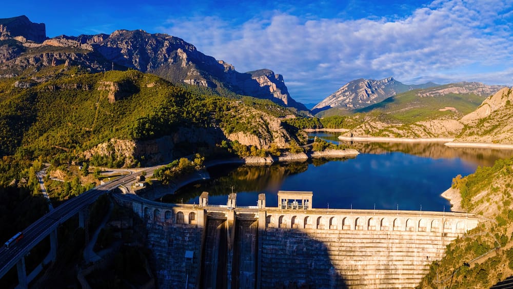 A dam with a reservoir, mountains, and a road.