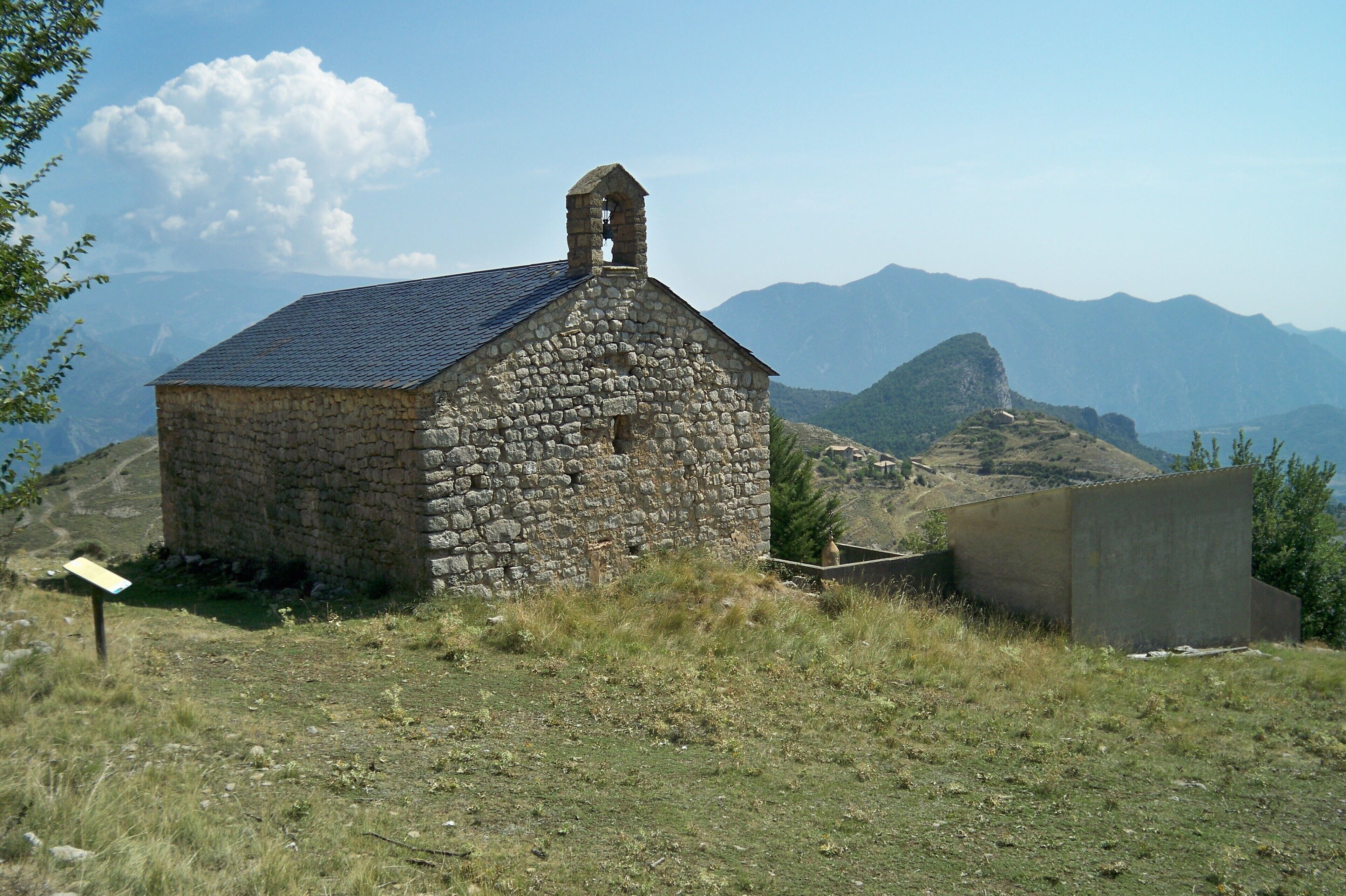 Vista del conjunt del recinte de l'església parroquial de Sant Joan de Montanissell des d'on sobserven les façanes nort i oest, els ninxos del cementiri i l'esglesia de la Mare de Déu de la Salut al turó dels fons. S'observa perfectament la pissarra de la teulada fruit de la seva reparació en temps recents.