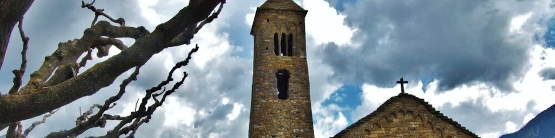 A stone church with a bell tower and a cross on top, surrounded by mountains and a cloudy sky.