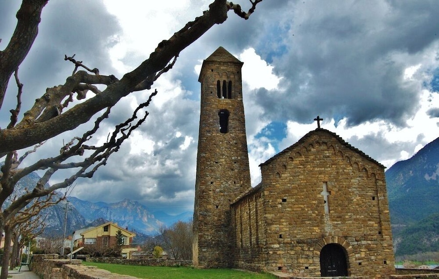 Una iglesia de piedra con un campanario y una cruz en la parte superior, rodeada de montañas y un cielo nublado.