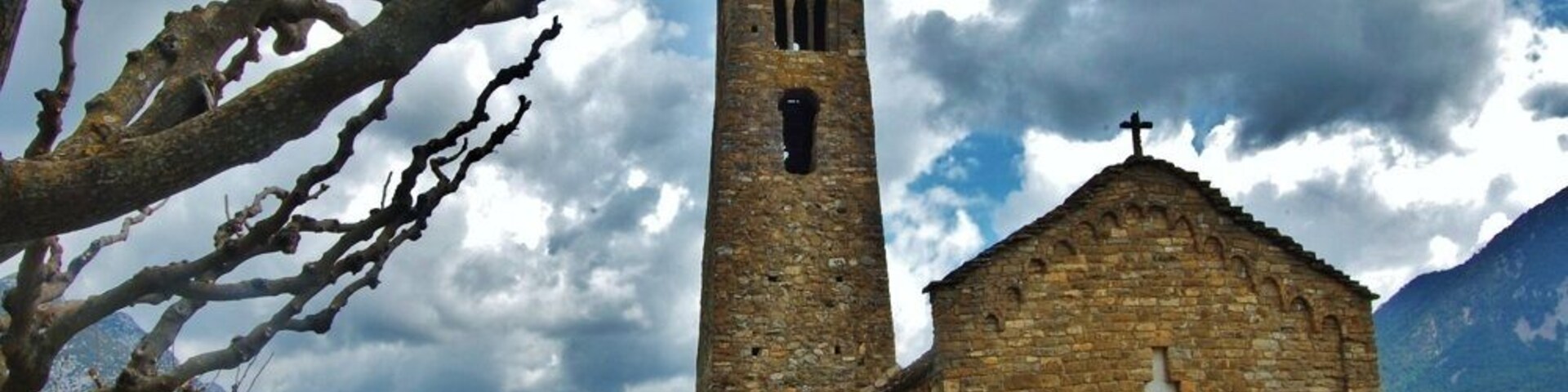 Uma igreja de pedra com uma torre de sino e uma cruz no topo, rodeada por montanhas e um céu nublado.