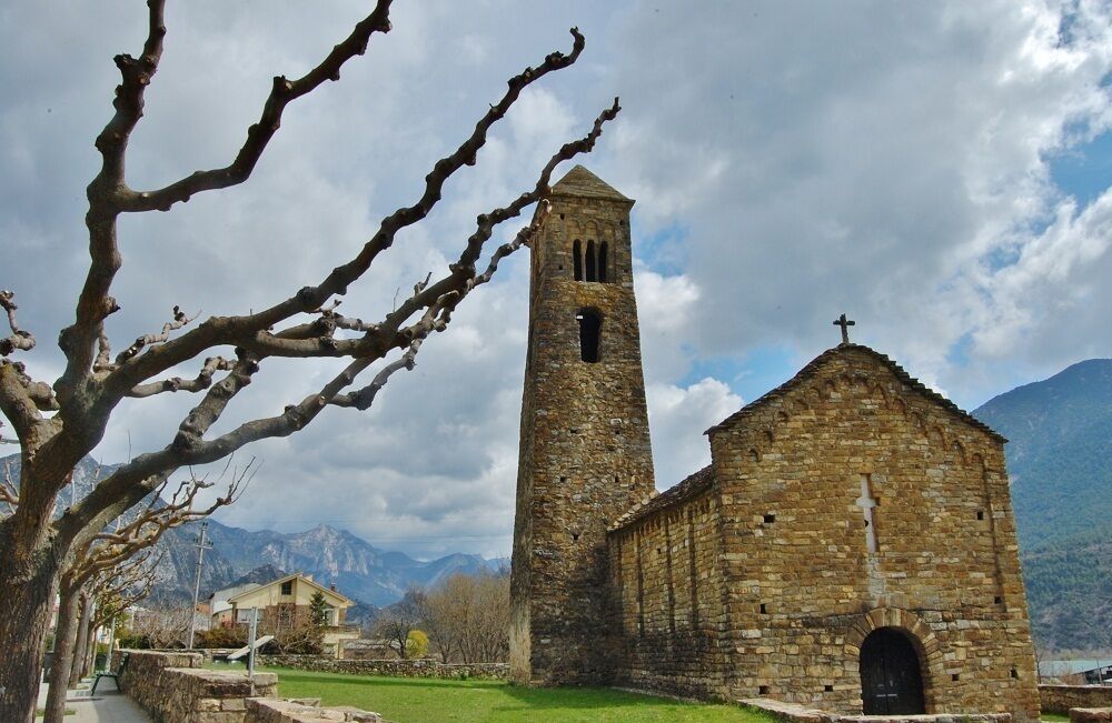 A stone church with a bell tower and a cross on top, surrounded by mountains and a leafless tree.