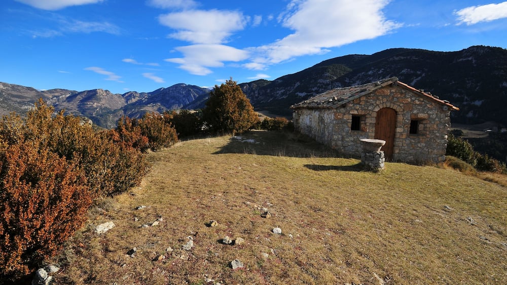 Ein Steinhhaus mit Holztür, umgeben von Herbstlaub und Bergen im Hintergrund.