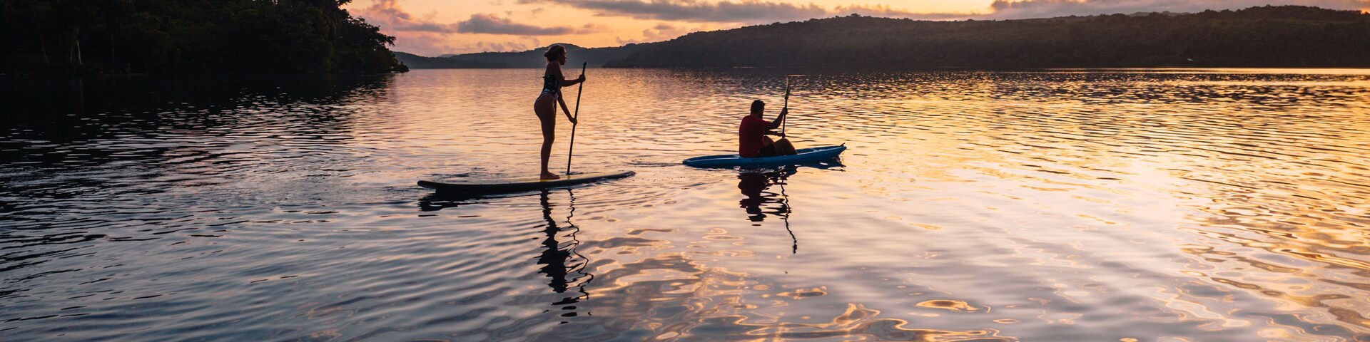 Una puesta de sol sobre un lago tranquilo con dos personas practicando remo de pie.
