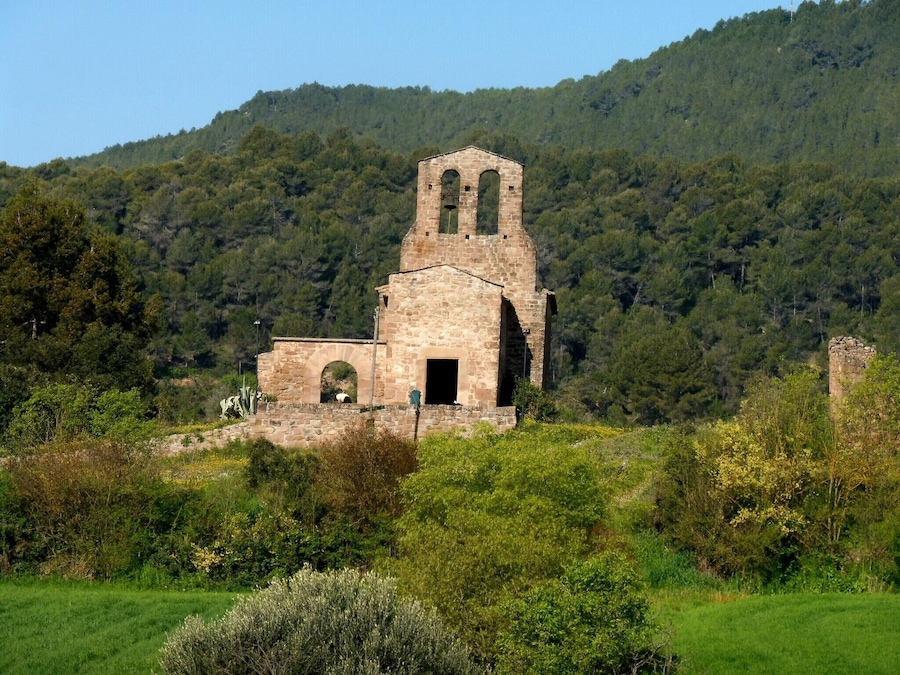 Una iglesia de piedra antigua con ventanas de arco apuntado y un campanario, rodeada de vegetación exuberante y una colina boscosa al fondo.
