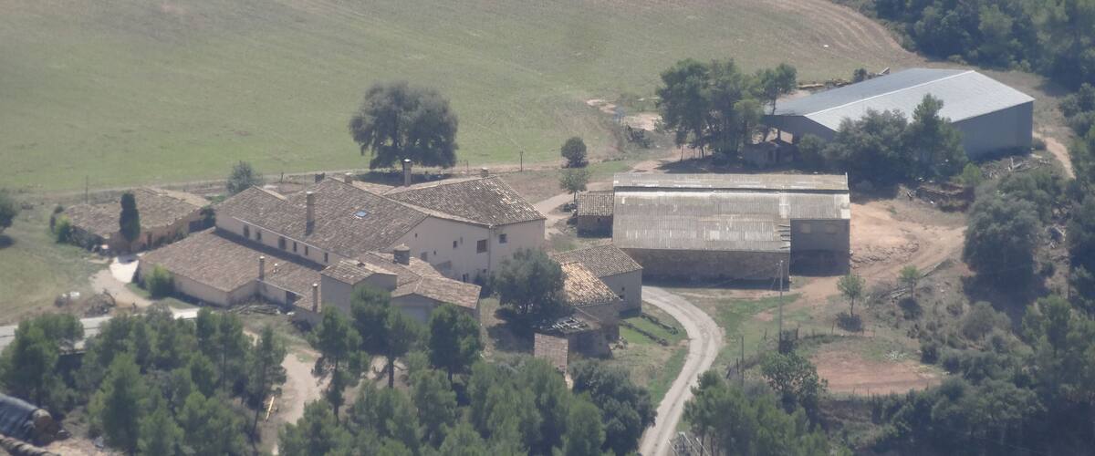An aerial view of a rural area with a large barn, several smaller buildings, a winding road, and a dense forest in the background.
