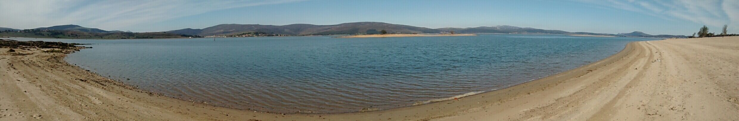 Een breed zandstrand met kalm water en heuvels in de verte.