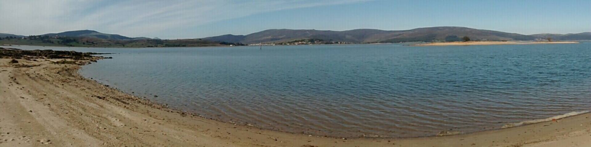 A wide sandy beach with calm water and distant hills.