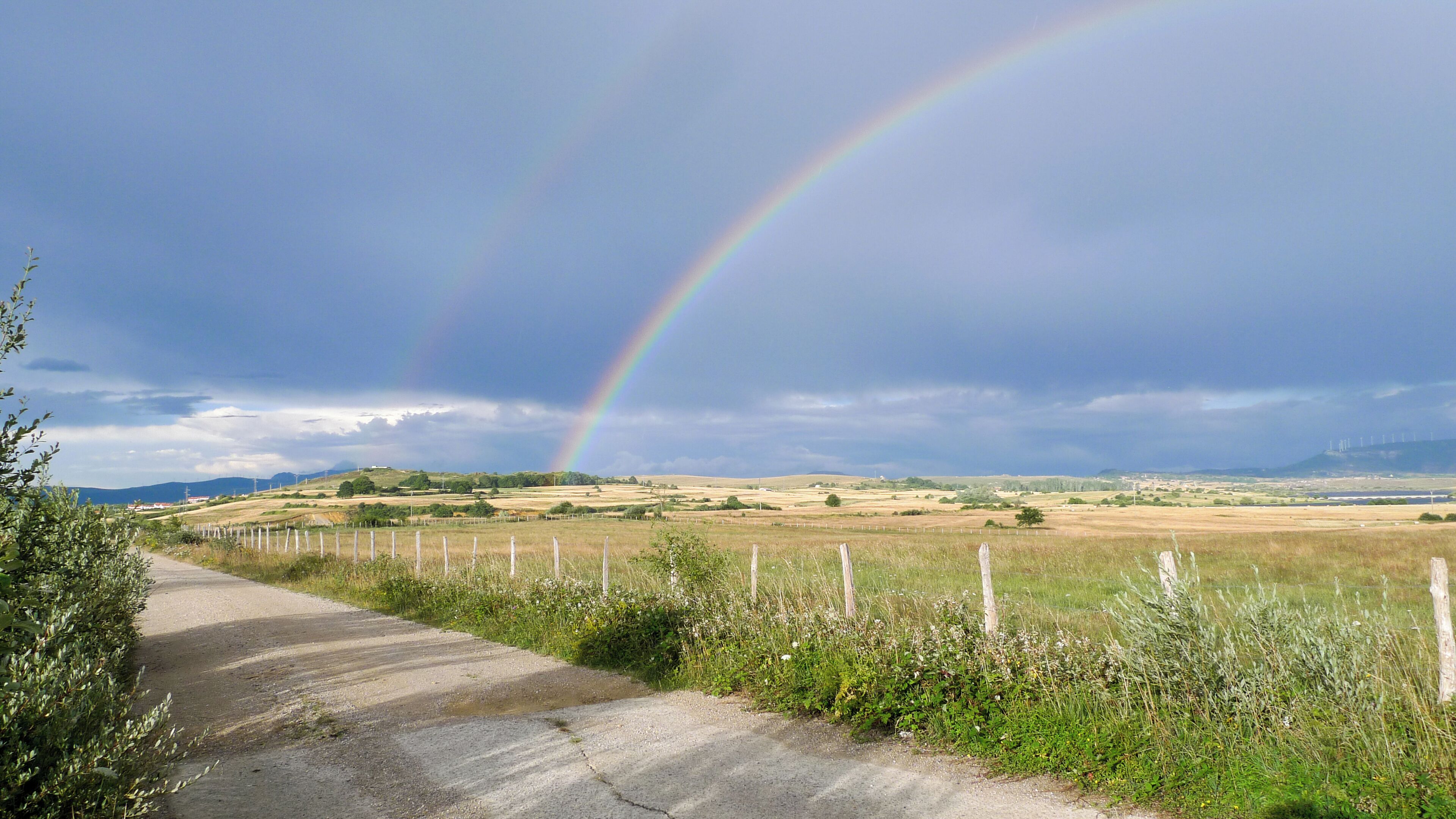 A rural landscape with a dirt road, grassy fields, and a rainbow.