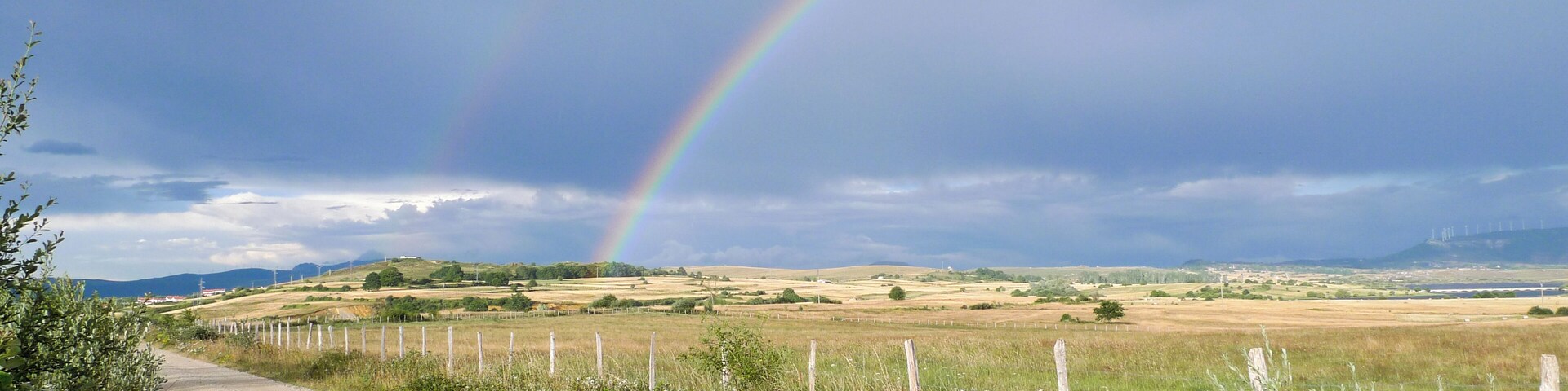 A rural landscape with a dirt road, grassy fields, and a rainbow.