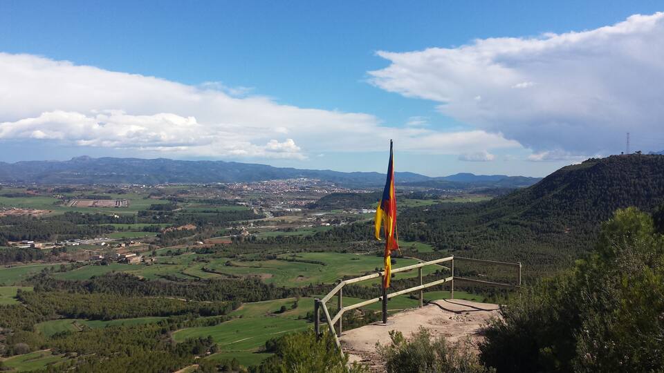 A flagpole with a flag, a mountainous landscape, and a valley with scattered buildings.
