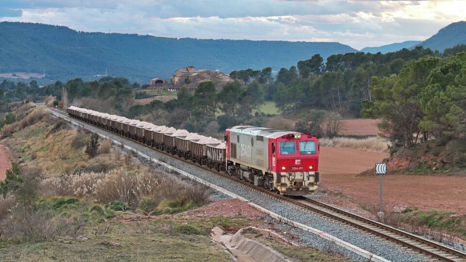 A red train traveling through a rural landscape with hills and trees in the background.