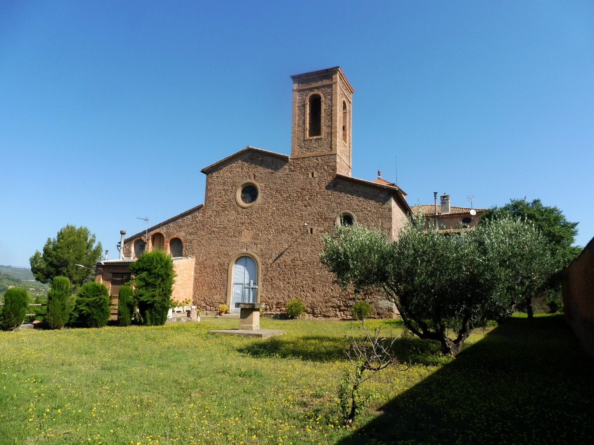 A historic stone church with a bell tower, surrounded by a grassy area and trees.