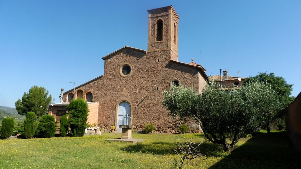 A historic stone church with a bell tower, surrounded by a grassy area and trees.