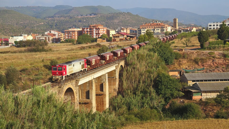 A train crossing a stone bridge over a river, with buildings and hills in the background.