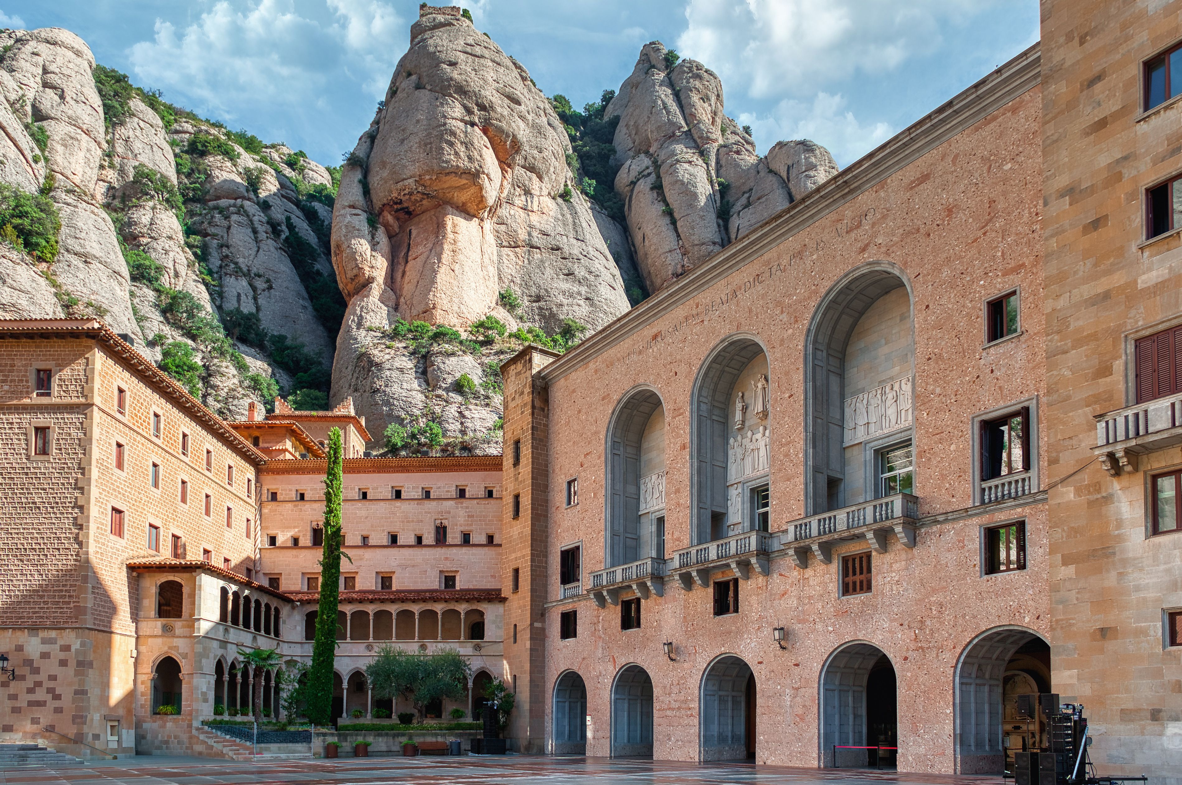 Um pátio com uma grande formação rochosa, um edifício com arcos e montanhas ao fundo.