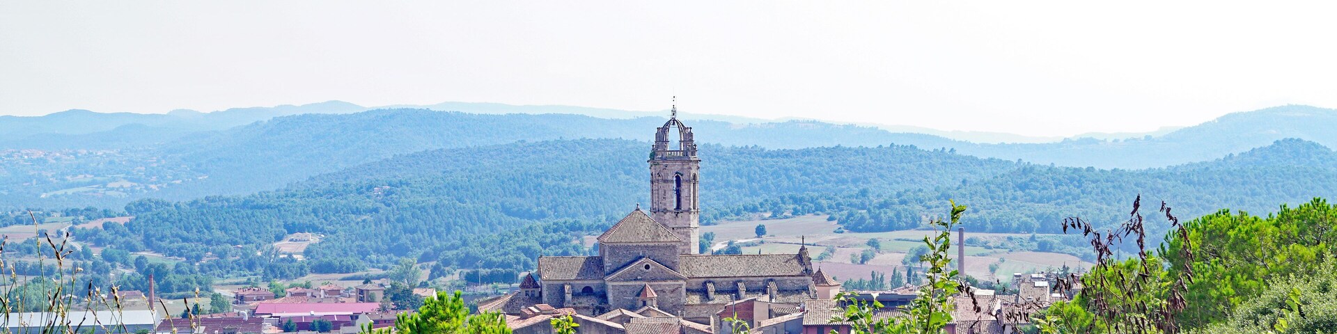 A historic church with a tall bell tower surrounded by lush greenery and rolling hills in the background.