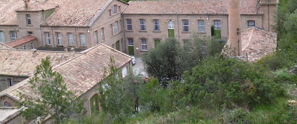 A historic building with a tall chimney, surrounded by lush greenery and a hilly landscape.