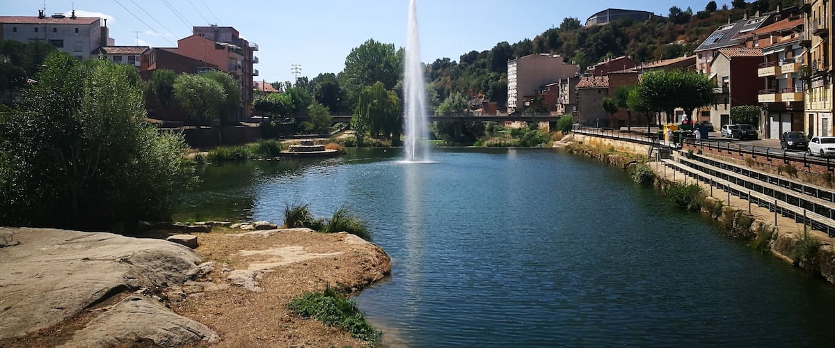 A river with a central fountain, surrounded by buildings and greenery.
