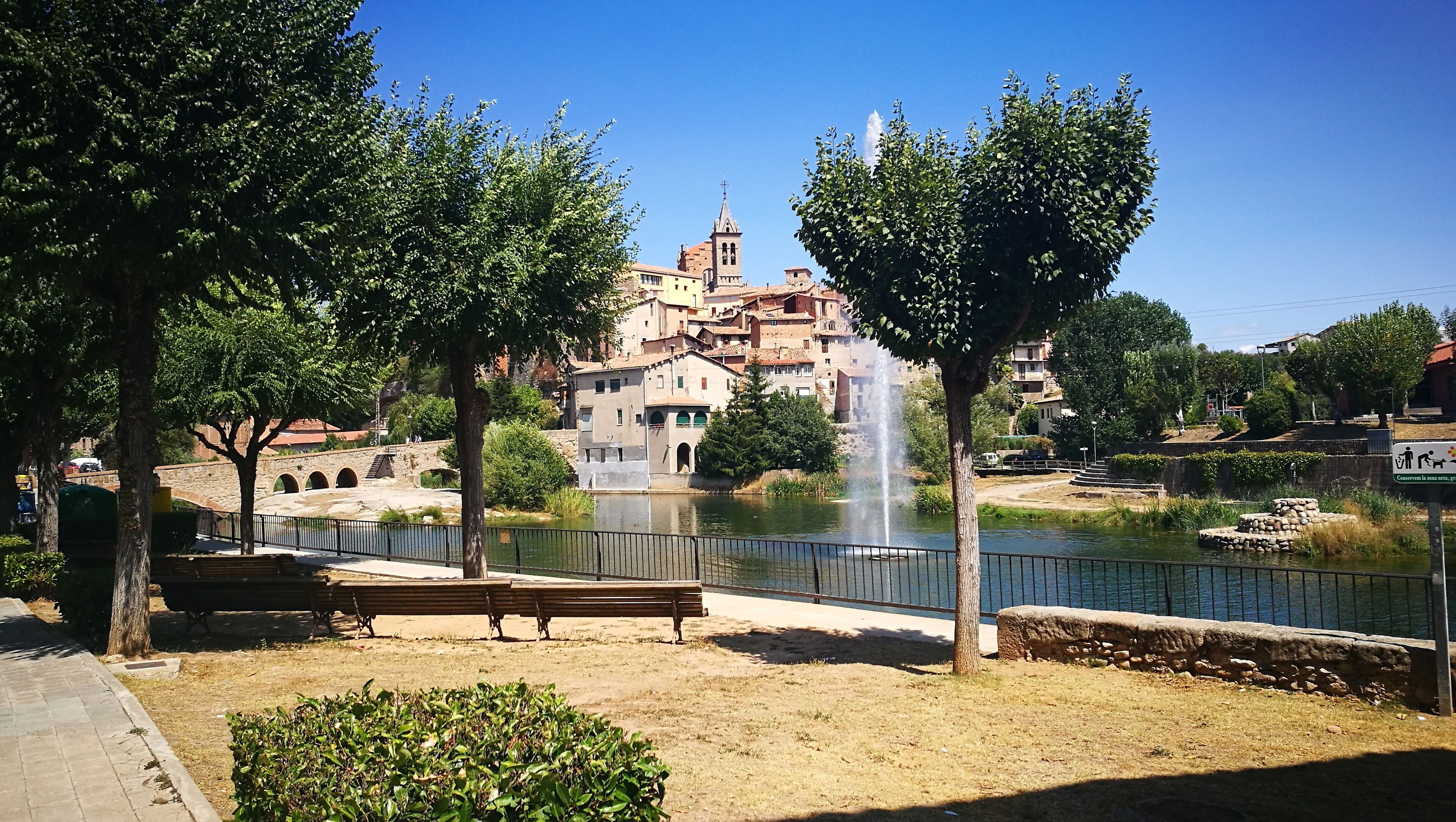 A park with a fountain, benches, trees, and a historic building with a tower in the background.