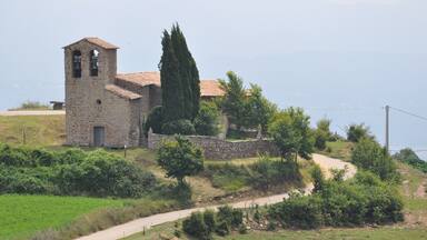 A stone church with a bell tower, surrounded by greenery and a dirt path.