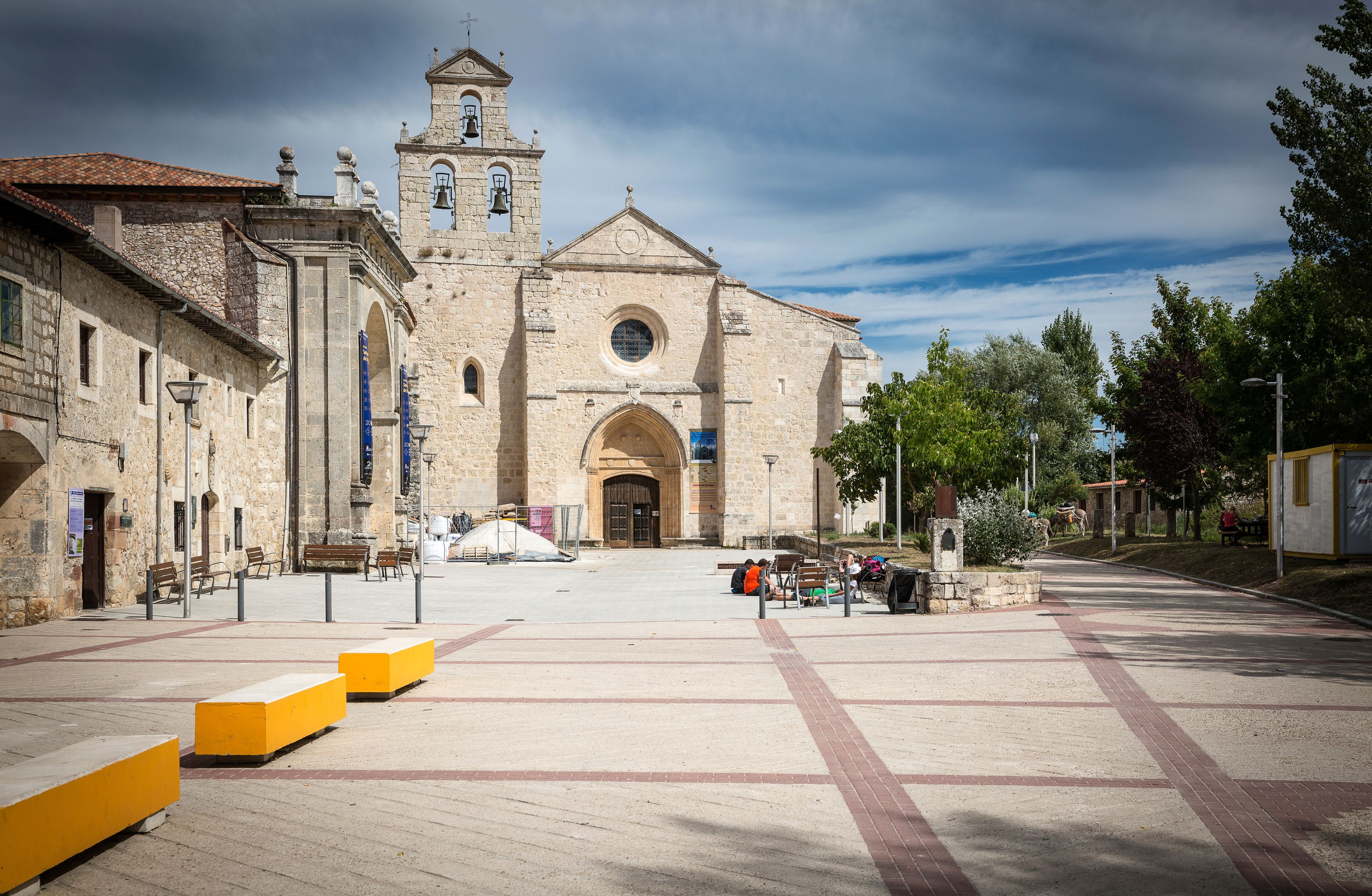 A historic church with a bell tower, a paved square with benches, and surrounding trees.