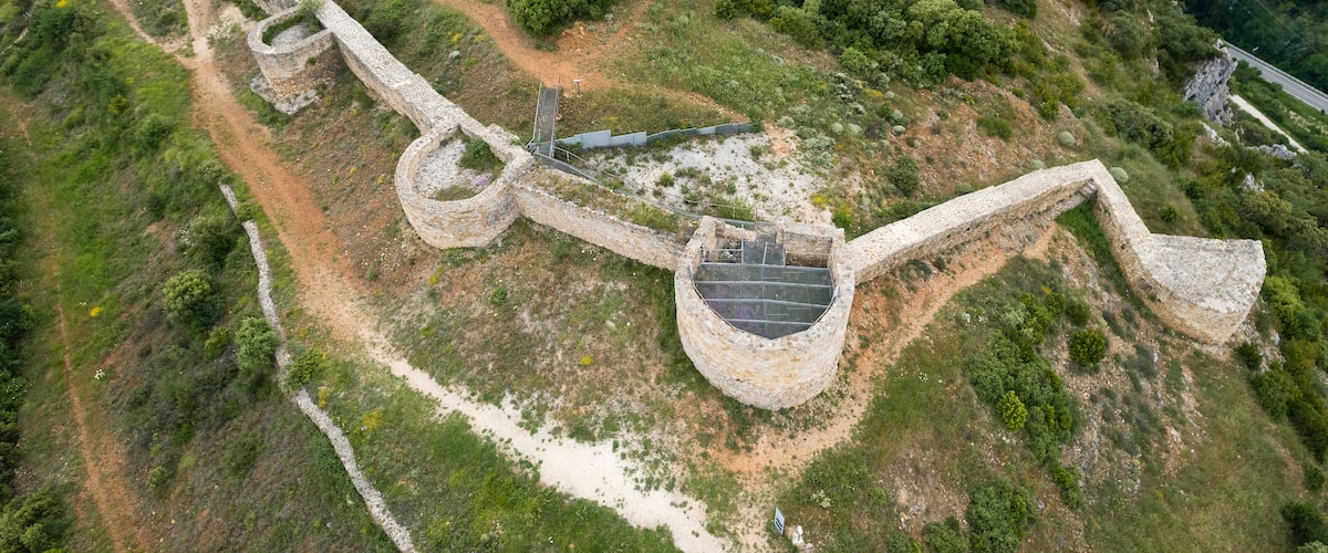 An aerial view of a historical stone fortification with a winding path, surrounded by dense forest and hills.