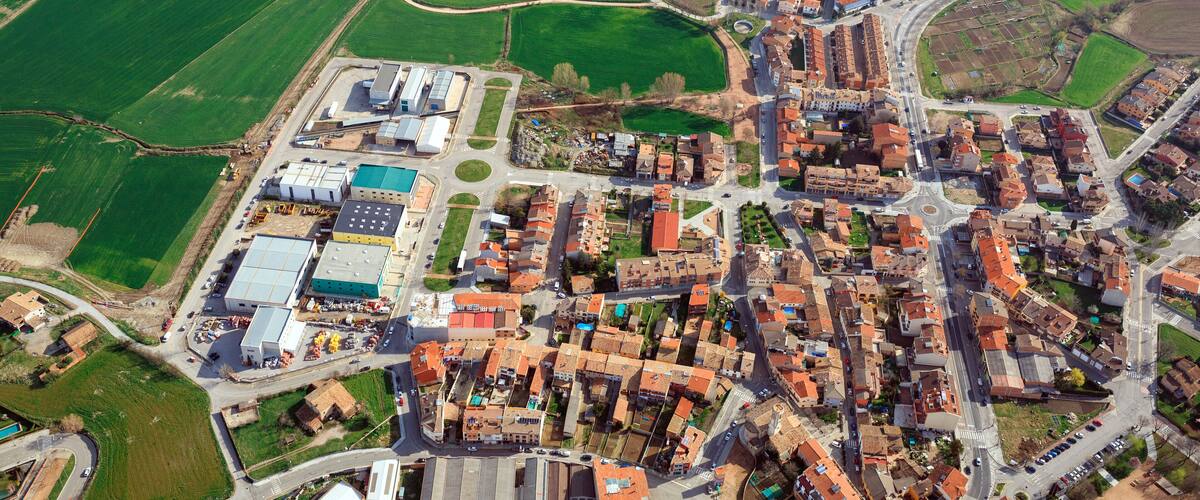 An aerial view of a town with a mix of residential and industrial areas, surrounded by green fields and a large body of water.