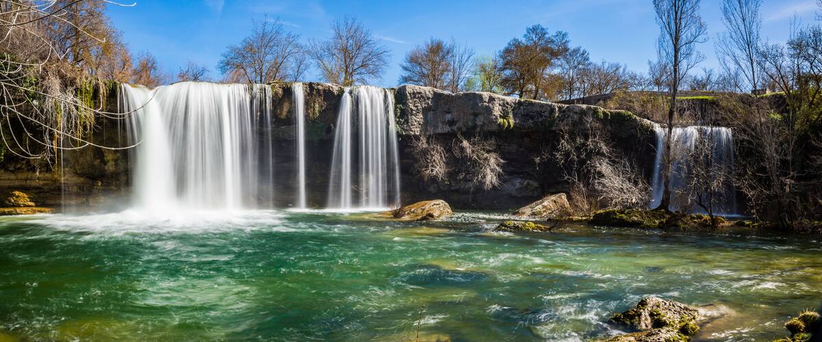 A waterfall cascading over rocks into a clear river surrounded by trees.