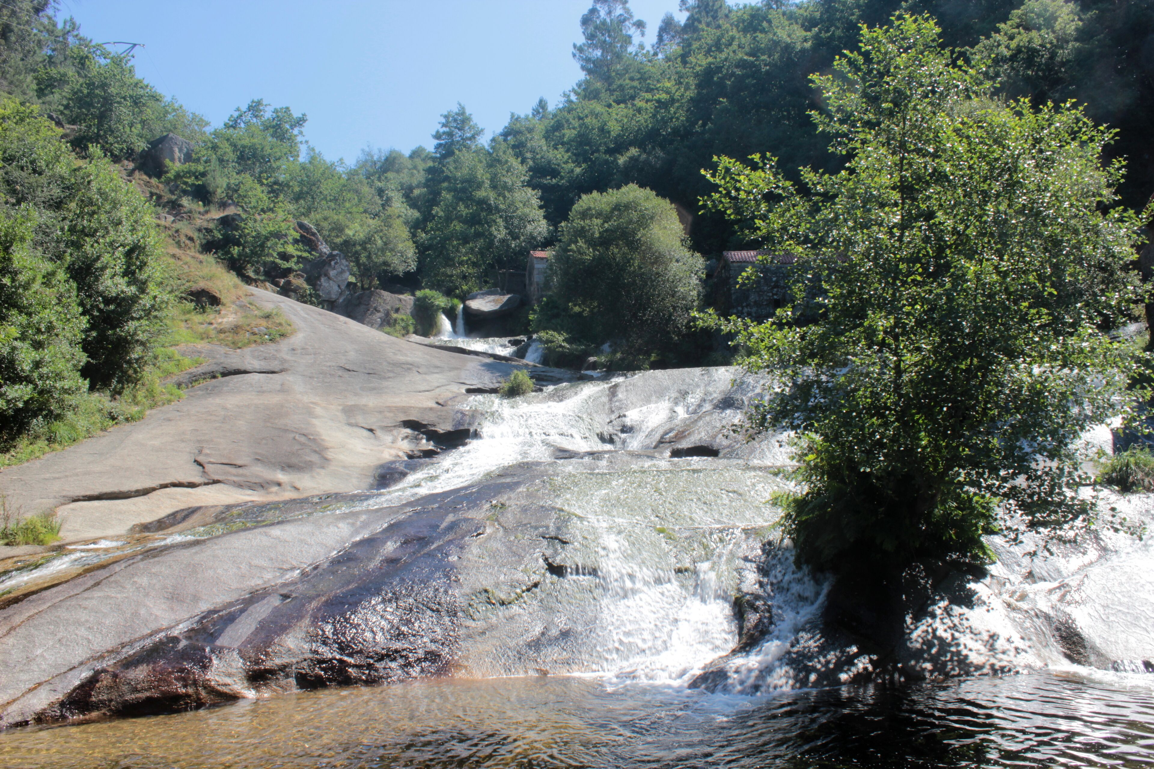 A rocky waterfall in a forested area with trees and a clear sky.
