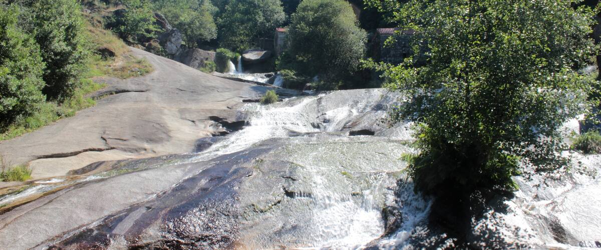 A rocky waterfall in a forested area with trees and a clear sky.