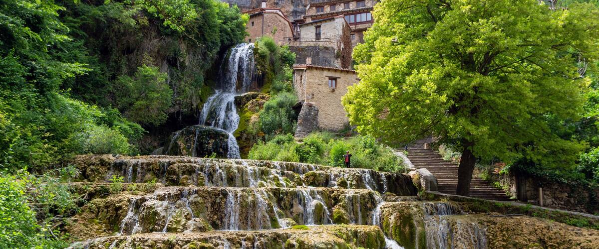 A multi-tiered waterfall surrounded by lush greenery and traditional stone buildings.