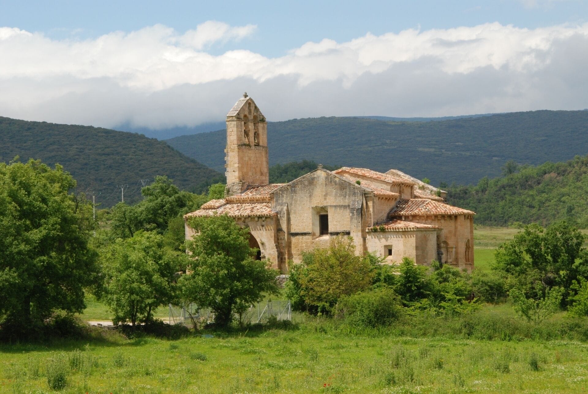 An old stone church with a bell tower surrounded by lush greenery and hills in the background.