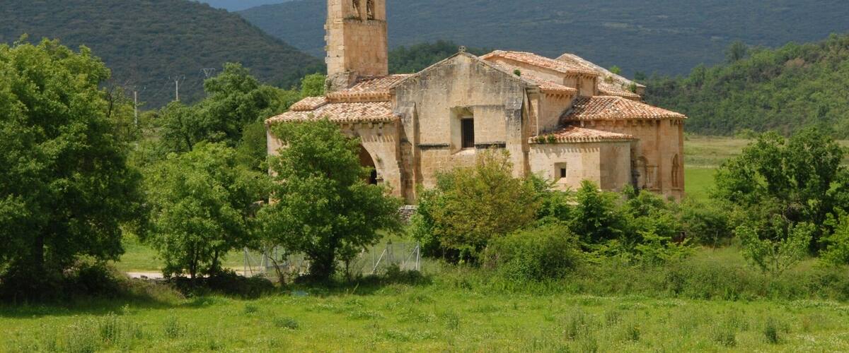 An old stone church with a bell tower surrounded by lush greenery and hills in the background.
