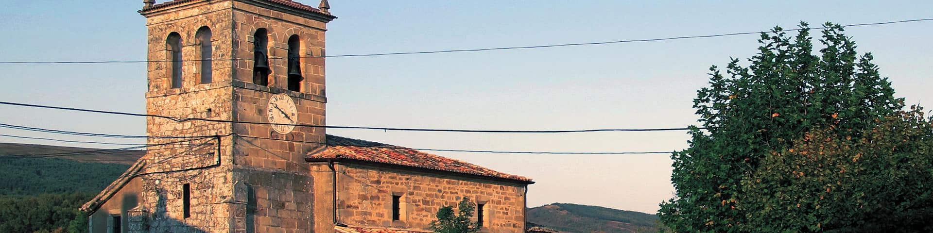 An old stone church with a bell tower and a clock, surrounded by trees and a road.