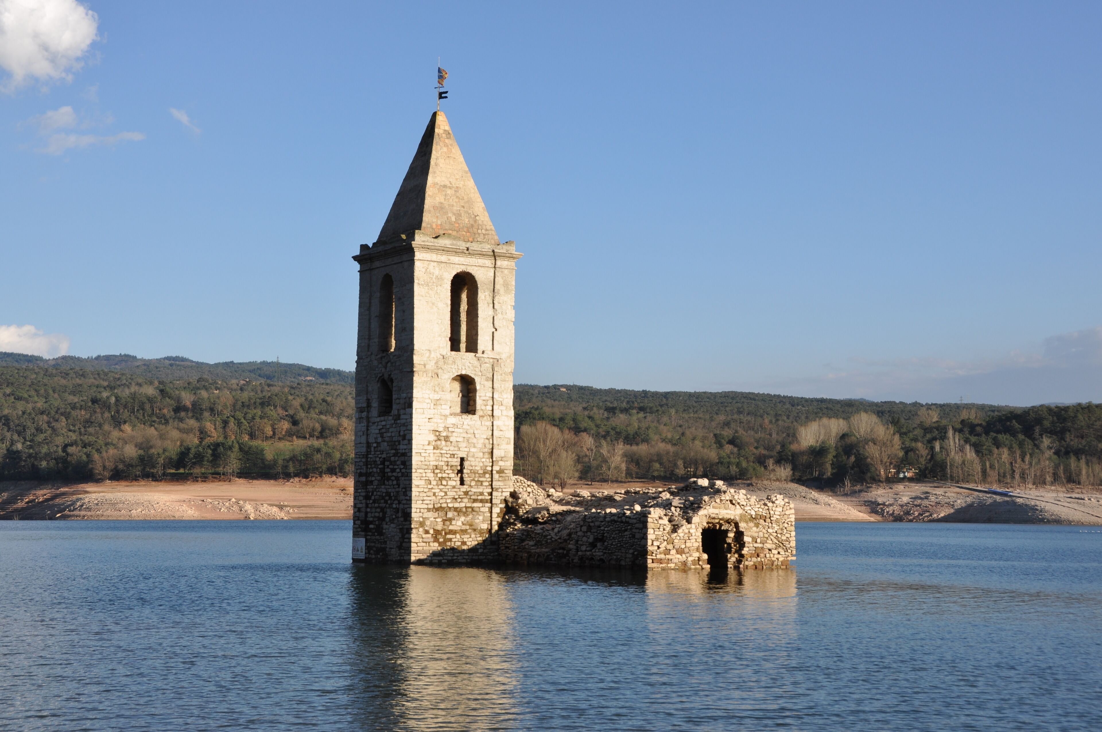 A stone tower by a calm lake with a forested hillside in the background.