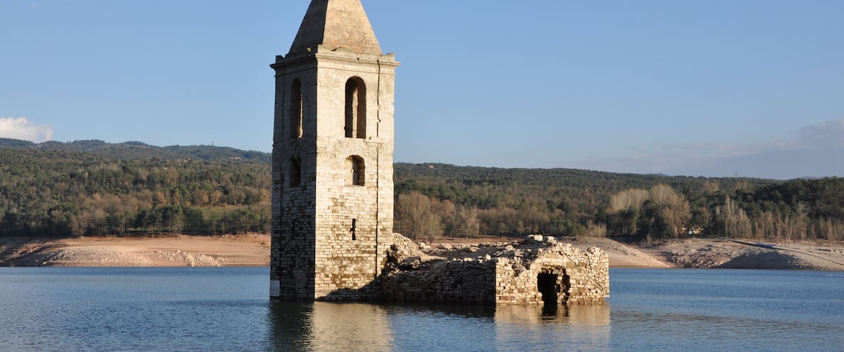 A stone tower by a calm lake with a forested hillside in the background.
