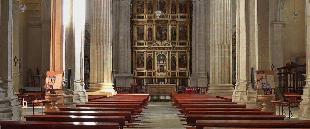 A church interior with rows of wooden pews, stone columns, and a large altar with religious paintings.