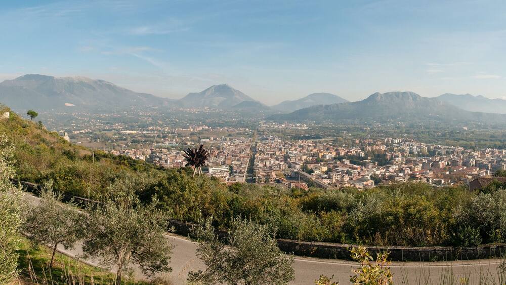 Un paysage urbain dominé par une colline sur laquelle se dresse une structure ressemblant à un château, le tout étant entouré d’une végétation luxuriante et avec des montagnes en arrière-plan.