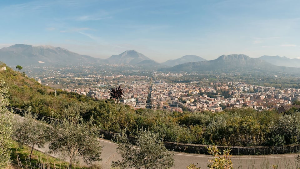 A cityscape with a prominent hill featuring a castle-like structure, surrounded by lush greenery and mountains in the background.