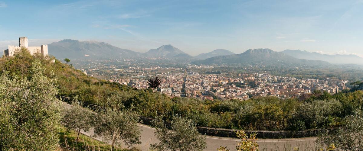 A cityscape with a prominent hill featuring a castle-like structure, surrounded by lush greenery and mountains in the background.