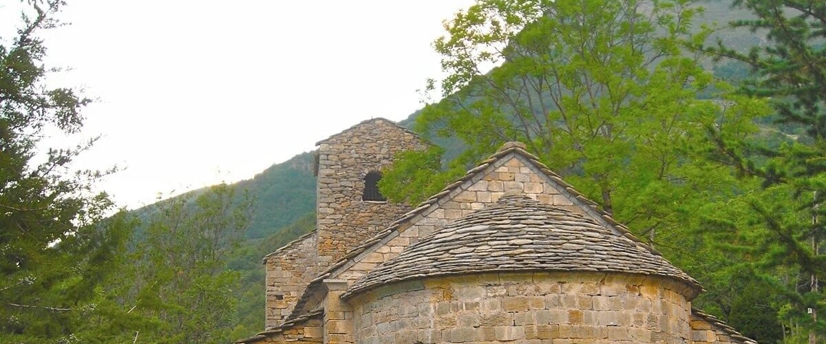 Una iglesia de piedra con un campanario, rodeada de abundante vegetación y colinas.
