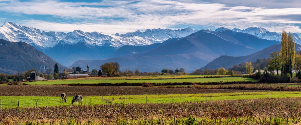 A mountainous landscape with snow-capped peaks, green fields, and a few buildings in the foreground.