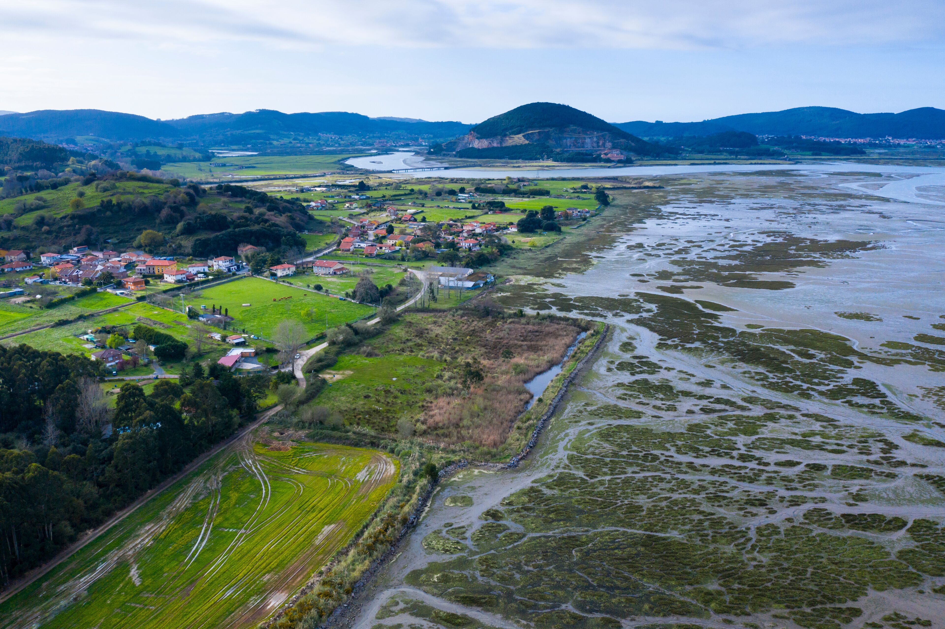 An aerial view of a coastal town with a river, green fields, and a hilly landscape in the background.