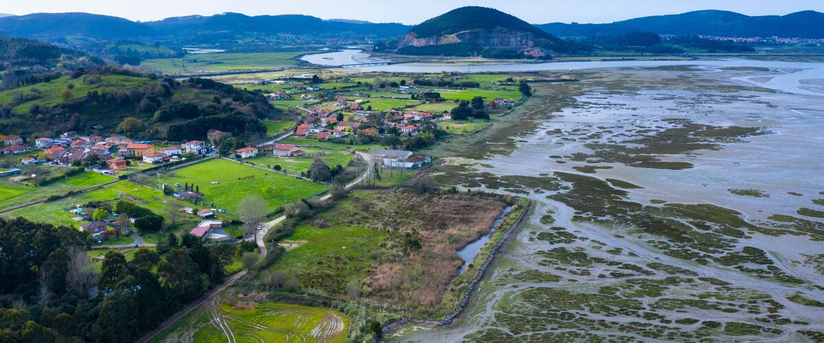 An aerial view of a coastal town with a river, green fields, and a hilly landscape in the background.