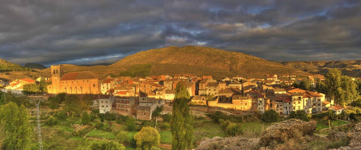 A town with historic buildings and a prominent church, surrounded by hills and a cloudy sky.
