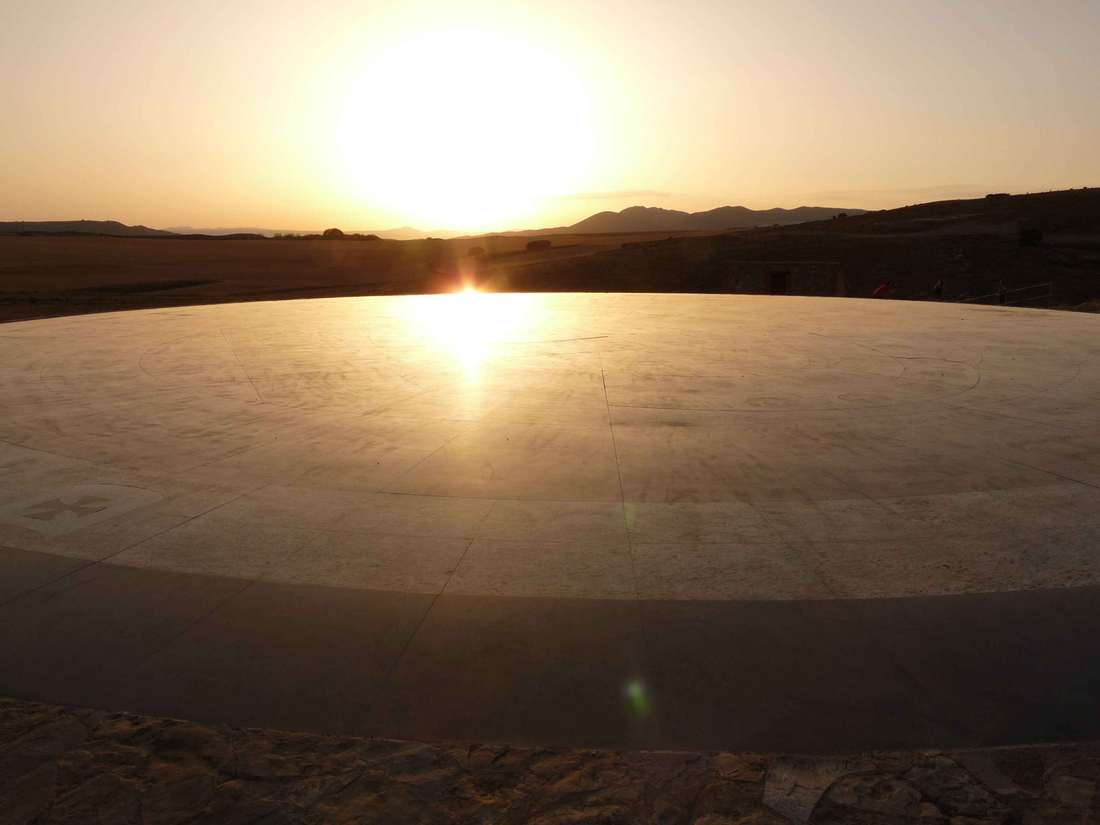 A circular outdoor area with a flat surface, surrounded by a rocky landscape and distant hills.