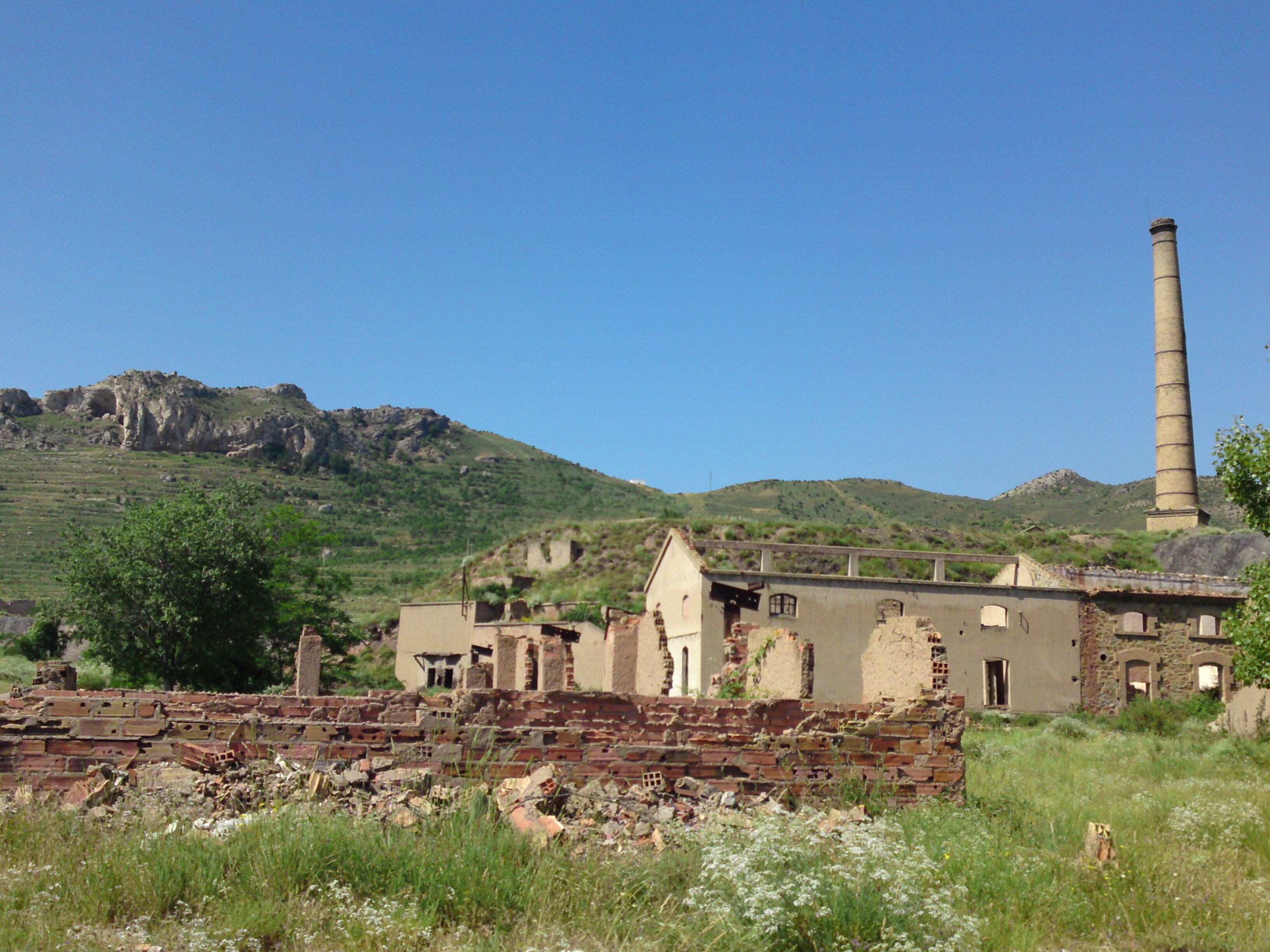 A dilapidated industrial site with a chimney, scattered ruins, and a mountainous backdrop.